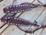 A pair of intricately tooled leather horse tack pieces, designed in a belt style headstall with Western motifs, featuring leaf patterns and white accents. Resting on a wooden surface, they include silver buckle detailing on one end. The product is the White Buckstitch Browband Headstall.