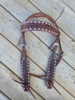 A detailed, light-brown White Buckstitch Browband Headstall with white stitching and intricate carvings laid out on a wooden deck. The bridle features two silver buckles for adjustments and leather ties at the ends.