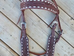 A detailed, light-brown White Buckstitch Browband Headstall with white stitching and intricate carvings laid out on a wooden deck. The bridle features two silver buckles for adjustments and leather ties at the ends.