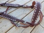 A White Buckstitch Browband Headstall lies on a wooden surface. The leather is intricately embossed with decorative patterns, highlighting the craftsmanship. Designed in a belt style headstall, it combines function and elegance perfectly.