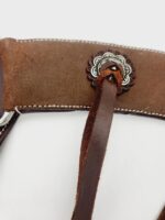 Close-up of a brown leather strap with white stitching, featuring a decorative concho with scalloped edges in the center. A leather lace is looped through the concho. The background is plain white.