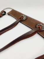 Close-up of a brown leather strap with intricate metal embellishments and multiple thinner leather strips looped through it. The strap has a silver ring at one end. The background is plain white.