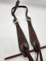 A brown leather horse bridle with metal buckles and decorative silver embellishments is laid out on a white surface. The bridle features white stitching along the edges, and leather straps extending from each end.