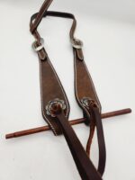 A brown leather horse breast collar with intricate silver buckles and floral conchos is laid out on a white surface. The collar features a t-strap and detailed stitching along the edges, showing a rustic and sturdy design for equestrian use.