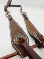 Close-up of brown leather horse bridle with intricate silver embellishments, white stitching, and metal buckles. It is draped on a wooden rod, showcasing the craftsmanship and details of the leather work against a plain white background.