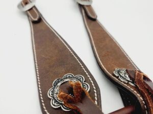 Close-up of brown leather horse bridle with intricate silver embellishments, white stitching, and metal buckles. It is draped on a wooden rod, showcasing the craftsmanship and details of the leather work against a plain white background.