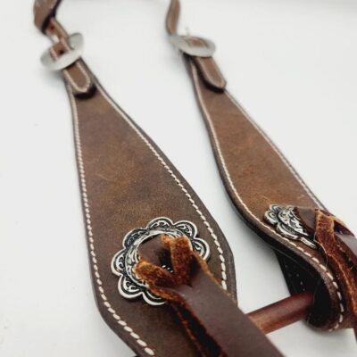 Close-up of brown leather horse bridle with intricate silver embellishments, white stitching, and metal buckles. It is draped on a wooden rod, showcasing the craftsmanship and details of the leather work against a plain white background.