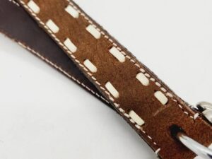 Close-up image of a brown leather strap with white stitching and woven detailing. The strap appears to be part of an accessory, possibly a bag or a piece of equipment. A metal ring is attached to one end of the strap. The background is plain white.