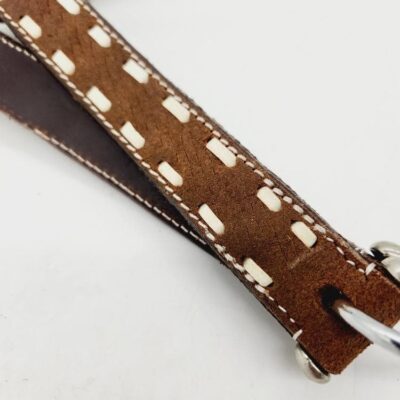 Close-up image of a brown leather strap with white stitching and woven detailing. The strap appears to be part of an accessory, possibly a bag or a piece of equipment. A metal ring is attached to one end of the strap. The background is plain white.