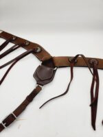 A close-up of a brown leather horse bridle with intricate stitching. The bridle features metal loops and leather straps with some hanging loosely. The background is plain white, highlighting the rich texture and craftsmanship of the leather.