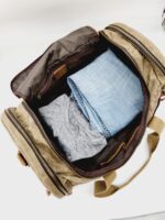 A brown duffel bag is shown open with visible contents including a neatly folded pair of light blue jeans and a gray t-shirt. The bag has multiple compartments and tan straps. The background is white and minimalistic.