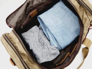 A brown duffel bag is shown open with visible contents including a neatly folded pair of light blue jeans and a gray t-shirt. The bag has multiple compartments and tan straps. The background is white and minimalistic.