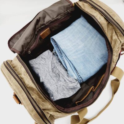 A brown duffel bag is shown open with visible contents including a neatly folded pair of light blue jeans and a gray t-shirt. The bag has multiple compartments and tan straps. The background is white and minimalistic.