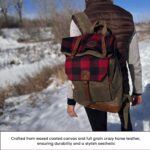 A person walks through a snowy landscape carrying a waxed coated canvas and full grain crazy horse leather backpack. The backpack features a red and black plaid blanket rolled up on top. The person is dressed warmly in a brown vest and gray long-sleeve shirt.