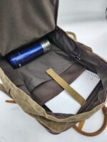 An open brown leather backpack reveals its contents: a blue thermos flask in a dedicated sleeve and a notebook in a compartment. The interior, lined with brown fabric, features a mustard-colored strap securing the notebook, embodying the rustic charm of The Rocky Mountain Zip Up Backpack- Explorer Series.