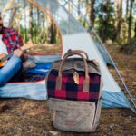 A plaid backpack is placed on the ground in front of a tent in a pine forest. In the background, a person in a red flannel shirt is sitting inside the tent, playing a ukulele.