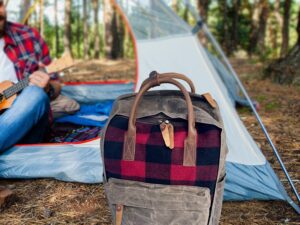 A plaid backpack is placed on the ground in front of a tent in a pine forest. In the background, a person in a red flannel shirt is sitting inside the tent, playing a ukulele.