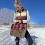 A woman stands on snow-covered ground under a clear blue sky, looking down at a plaid bag she is holding. She wears a striped, sleeveless winter vest over a gray long-sleeve shirt, black leggings, and has a braid over her shoulder. Leafless trees are in the background.