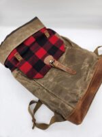 A vintage-style backpack with an olive green body, brown leather accents, and a red and black checkered flap secured by a leather strap and buckle. The backpack straps are also visible in the image.