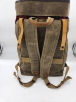 A weathered brown canvas backpack with leather accents on its adjustable straps is shown. The backpack has a top flap secured with leather straps and buckles. There are also side pockets and a handle at the top. The background is a plain white surface.