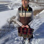 A person with a long braid stands on a snow-covered path, holding a plaid and leather satchel. They are wearing a color-blocked vest over a gray long-sleeve shirt, black pants, and snow boots. In the background, there are snow-covered trees and fields.