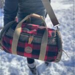 Person carrying a duffel bag with a red and black plaid pattern, leather straps, and a circular emblem. Snowy ground and winter attire in the background suggest a cold weather setting.