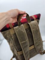 A hand is holding the top handle of a green canvas backpack with padded shoulder straps and a top section made of red and black plaid fabric. The background is out of focus.