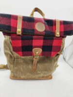 A brown and red plaid backpack with leather straps and accents. The top flap is red and black plaid with a leather patch in the center and snap closures. The front pocket is brown with a leather buckle strap. The bag has a carrying handle and adjustable shoulder straps.