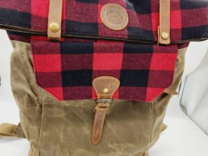 A brown and red plaid backpack with leather straps and accents. The top flap is red and black plaid with a leather patch in the center and snap closures. The front pocket is brown with a leather buckle strap. The bag has a carrying handle and adjustable shoulder straps.