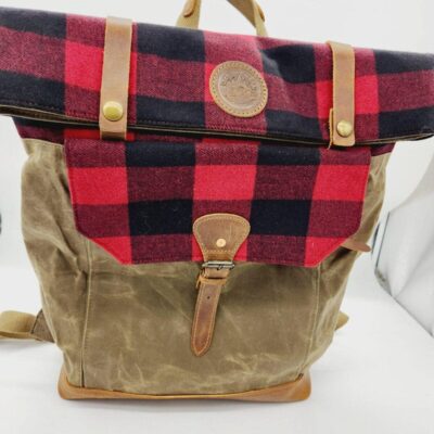A brown and red plaid backpack with leather straps and accents. The top flap is red and black plaid with a leather patch in the center and snap closures. The front pocket is brown with a leather buckle strap. The bag has a carrying handle and adjustable shoulder straps.