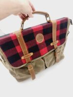 A hand holding a stylish messenger bag with a red and black plaid pattern on the flap, secured with brown leather straps and buckles. The main body of the bag is made from a durable, greenish-brown fabric with pockets on the front.