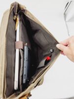 A hand holds open The Rocky Mountain Laptop Bag- Explorer Series, displaying its organized interior. Inside, there is a laptop secured by a strap, a black tablet, and a red item in a smaller pocket. The leather bag features multiple compartments and a zipper closure.