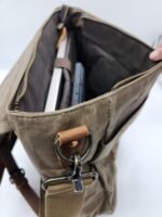 A brown fabric messenger bag with a leather strap is shown opened, revealing a laptop and another device inside. The Rocky Mountain Laptop Bag- Explorer Series, reminiscent of a leather saddle in its craftsmanship, has multiple compartments, metal clasps and buckles, and is partially held open by a hand at the top right corner.