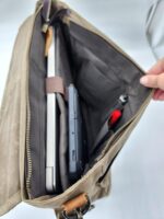 A hand holding open a beige messenger bag showing its contents. Inside are a laptop secured with a strap, a handheld gaming console, a red pen, and a small black item in one of the interior pockets. The Rocky Mountain Laptop Bag - Explorer Series boasts zippered closure and resembles the classic charm of a saddle blanket backpack.