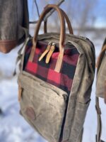 A close-up of a rugged The Rocky Mountain Zip Up Backpack - Explorer Series hanging on a branch. The backpack features a grey waxed canvas body with a front pocket adorned in red and black buffalo check fabric. Leather straps and zippers are seen, with a snowy landscape blurred in the background.