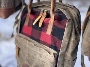 A close-up of a rugged The Rocky Mountain Zip Up Backpack - Explorer Series hanging on a branch. The backpack features a grey waxed canvas body with a front pocket adorned in red and black buffalo check fabric. Leather straps and zippers are seen, with a snowy landscape blurred in the background.