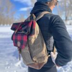 A person wearing a black jacket and a beanie stands in a snowy landscape, carrying a tan and red plaid backpack. They are wearing white gloves, and the background is filled with snow-covered trees and a clear blue sky.