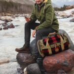 A person wearing a green jacket and brown boots sits on a rock by a river, holding a smartphone. A red and black plaid duffel bag is placed next to them. The background features mountains and a forest under overcast skies.