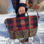 A person walks through a snowy landscape, holding a stylish bag adorned with a red and black buffalo plaid design and leather straps. The person's outfit includes a grey sweater layered under a brown jacket. The background is blurred, showing snow-covered ground and distant trees.