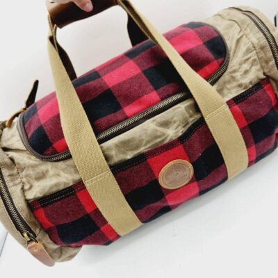 A person holding a cylindrical bag with a red and black plaid pattern and beige straps. The bag has multiple zippers and a round leather patch on the side. The background is white.