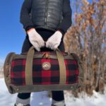 A person wearing a black puffer vest and white gloves holds a duffel bag with a red and black checkered pattern and tan straps. Snow covers the ground, and leafless trees are in the background under a bright blue sky.