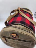 A cylindrical duffle bag with a combination of red and black plaid fabric and khaki canvas material. The bag features a front zippered pocket with a leather pull and two beige shoulder straps. The top of the bag also has a leather strap with a metal buckle.