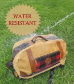 A tan and black duffel bag with leather accents and a red plaid pocket is shown with water being sprayed on it, demonstrating its water-resistant properties. The Rocky Mountain Perfect Duffel- Explorer Series, made from durable wax canvas, is on green grass, and the words "WATER RESISTANT" are prominently displayed above the bag.