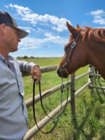 A person in a light-colored shirt and dark baseball cap stands beside a chestnut horse, holding its reins, with a wooden fence and green meadow in the background under a partly cloudy sky.