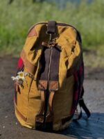 A worn yellow and brown backpack with leather straps sits on a dirt path. A small bouquet of wildflowers is tucked into the side pocket. The background is out of focus, displaying green foliage. The scene suggests a nature hike or outdoor adventure.