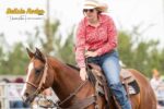 A person wearing a cowboy hat, red patterned shirt, and sunglasses rides a brown horse during the Delisle Rodeo. The rider is looking to the side with one hand on the reins and the other resting on the horse's neck. The background is blurred.