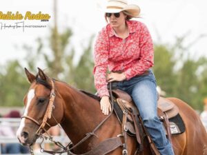 A person wearing a cowboy hat, red patterned shirt, and sunglasses rides a brown horse during the Delisle Rodeo. The rider is looking to the side with one hand on the reins and the other resting on the horse's neck. The background is blurred.