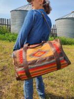 A person wearing a blue denim jacket and a hat is seen from behind, carrying a colorful, patterned duffel bag with leather straps over their shoulder. They are standing in a grassy area with silos and a fence visible in the background.