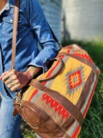 A person wearing a denim jacket and jeans is holding a large duffel bag with a colorful southwestern pattern and brown leather accents. The person's hand is tattooed, and they are walking outside on a sunny day. A corrugated metal wall and greenery are in the background.