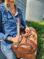 A person wearing a denim shirt and jeans, with tattoos visible on their neck and hands, is holding a large duffel bag. The bag features leather accents and a colorful, patterned fabric. The background includes a metal structure and green grass.